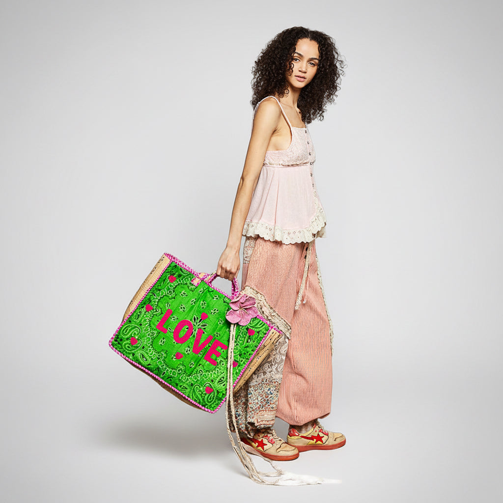 A woman stands against a white studio background holding a large green woven tote bag with pink lettering and a pink flower accent. She is wearing a pale pink ruffled camisole, loose pink corduroy pants, and sandals.