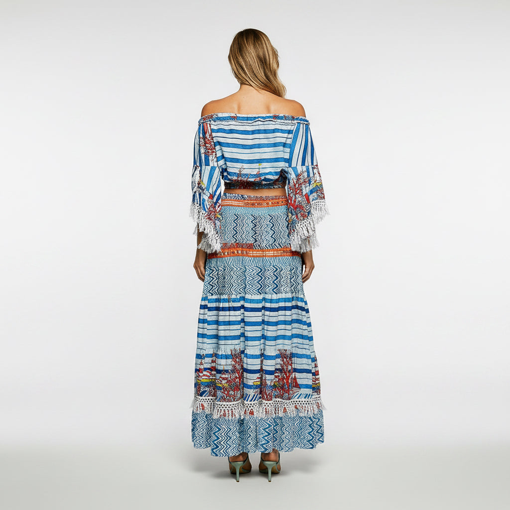 A woman viewed from the back wearing a long off-the-shoulder maxi dress with blue, white, and red geometric patterns and tassel trim, set against a plain white background.
