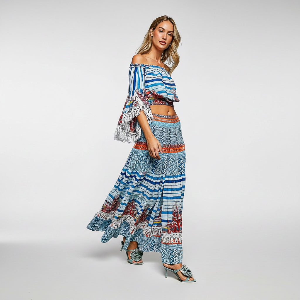 A woman modeling a two-piece bohemian-style maxi set featuring a blue, white, and orange patterned off-the-shoulder crop top with ruffled sleeves and a matching long skirt, set against a plain white background.