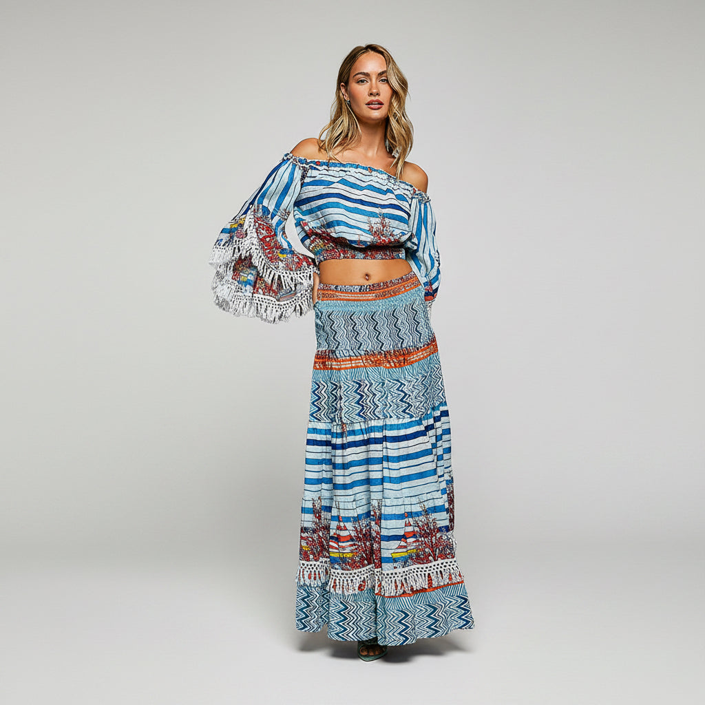 A woman modeling a bohemian-style two-piece set featuring a blue and white striped off-the-shoulder crop top with long fringed sleeves and a matching long skirt with geometric and floral patterns.
