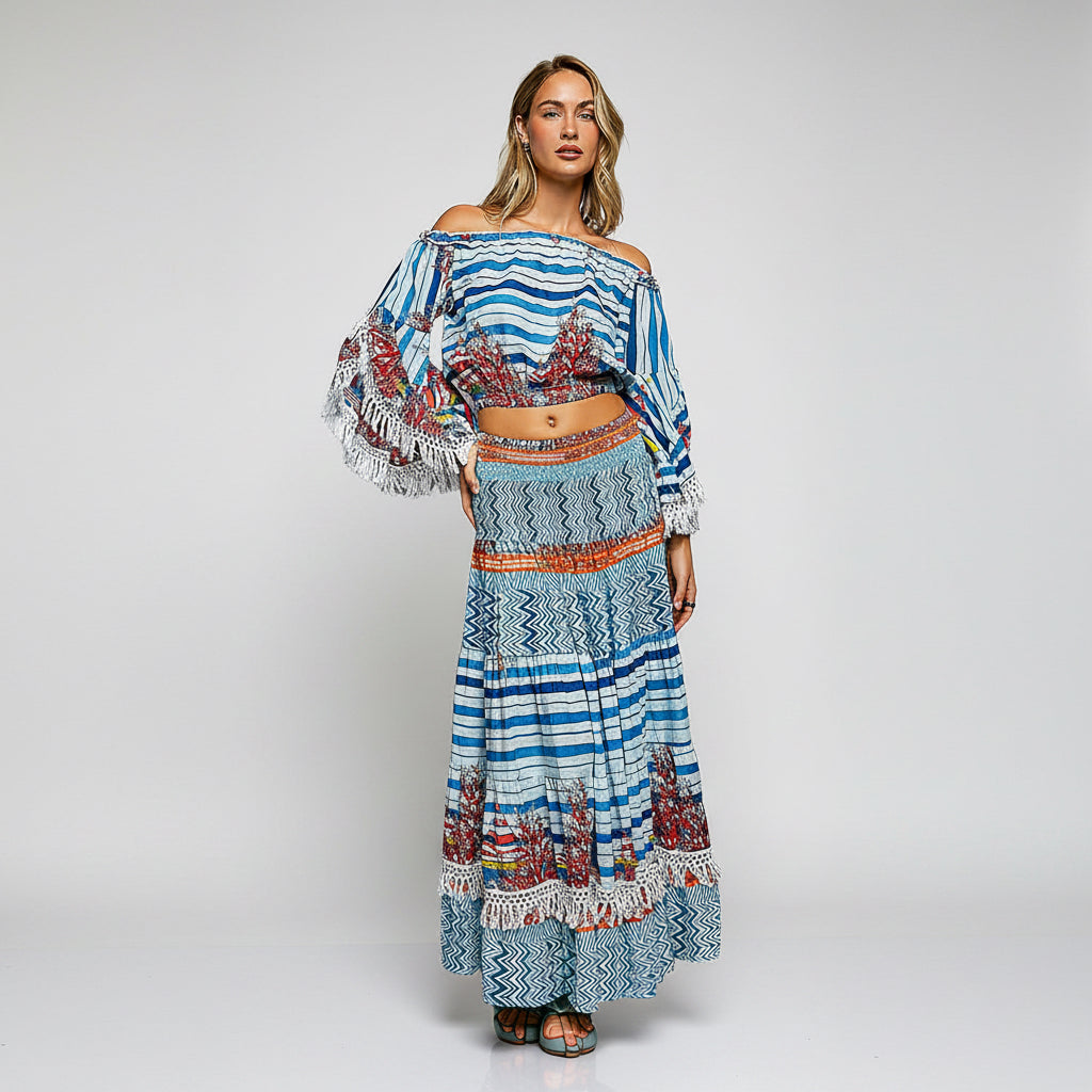 A woman modeling a two-piece bohemian-style outfit featuring a blue and white patterned crop top with long fringed sleeves and a matching high-waisted maxi skirt with geometric and tribal patterns.