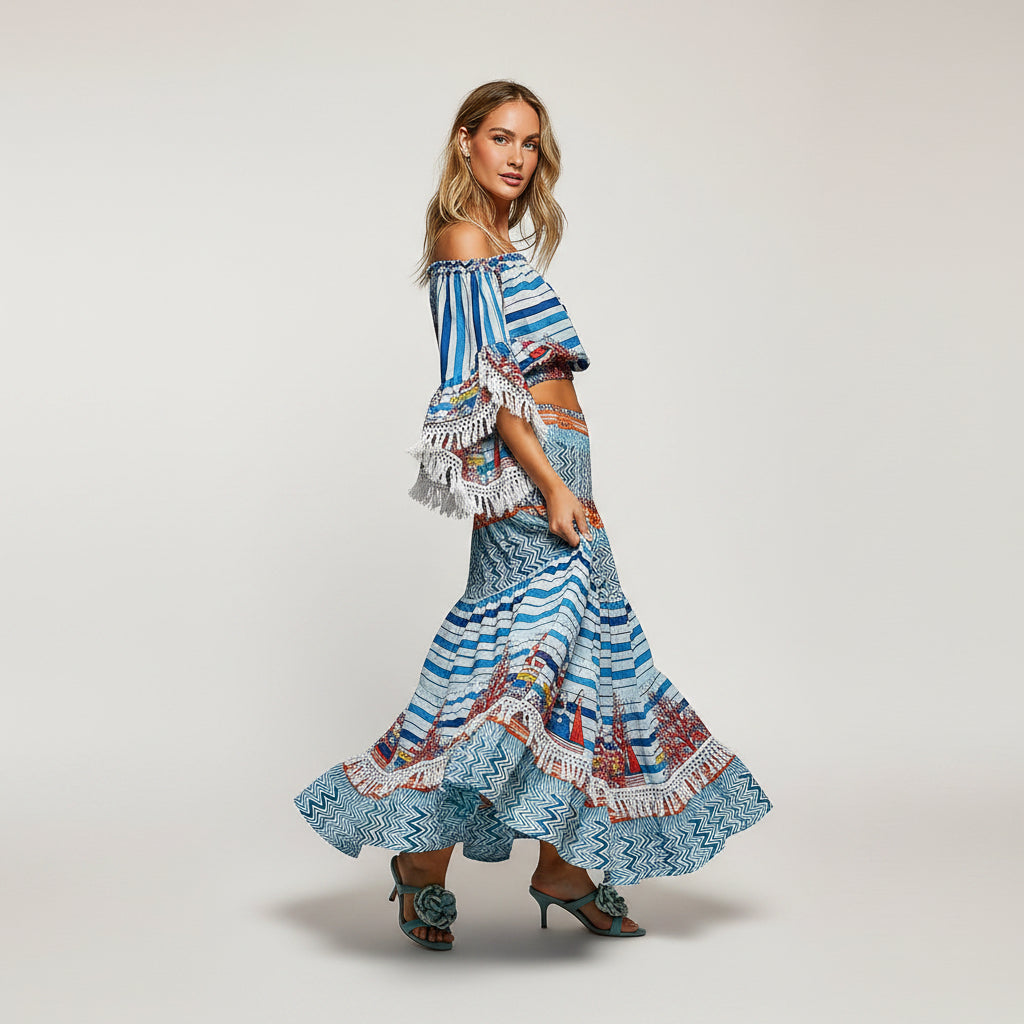 A woman modeling a two-piece bohemian-style maxi set featuring a blue and white patterned off-the-shoulder crop top with fringe and a matching tiered ruffled maxi skirt.