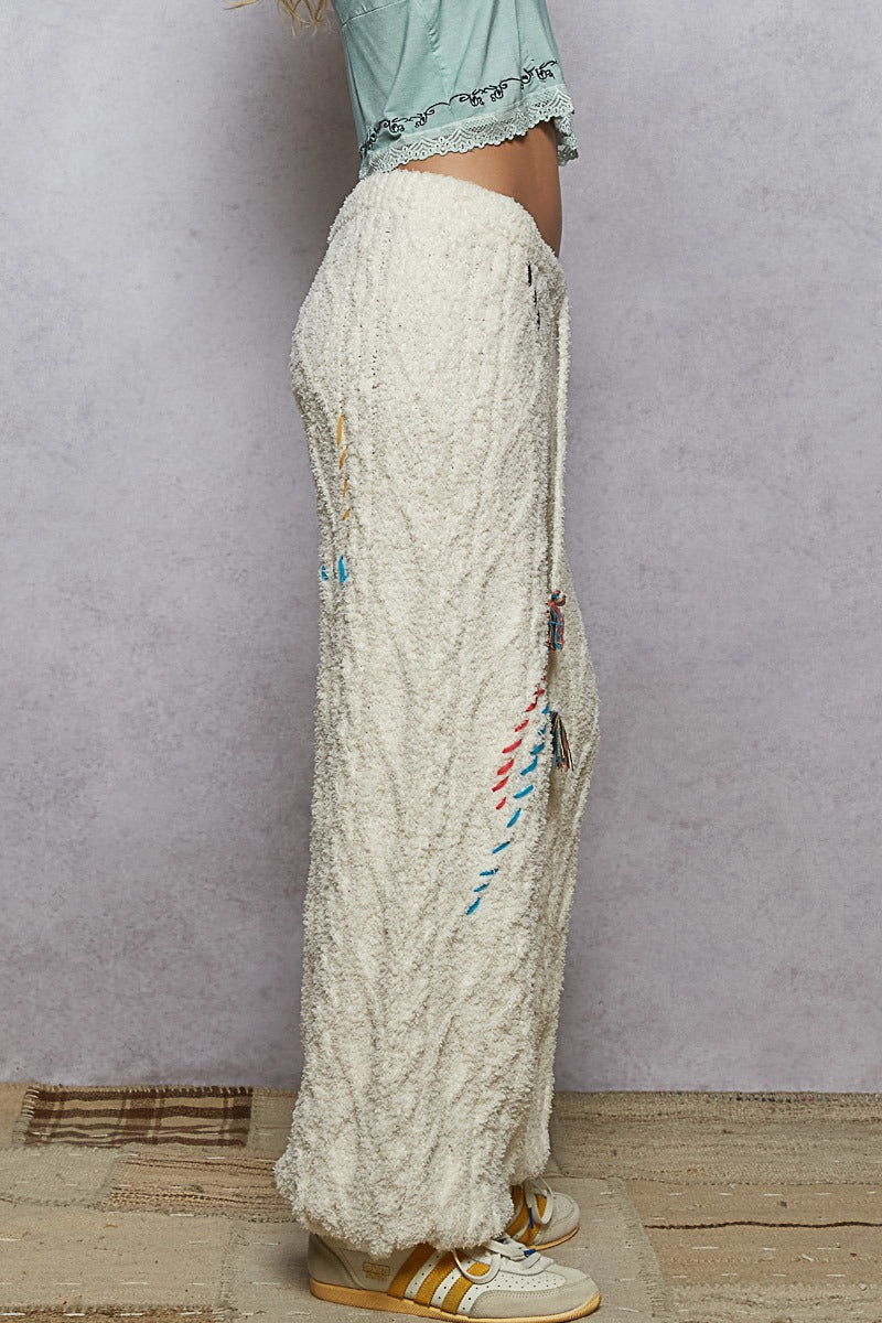 Person wearing white embroidered pants with colorful details on a plain background