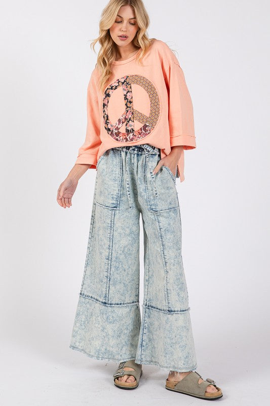 Woman wearing a peach-colored sweatshirt with a peace sign design and light blue denim wide-leg pants on a white background.