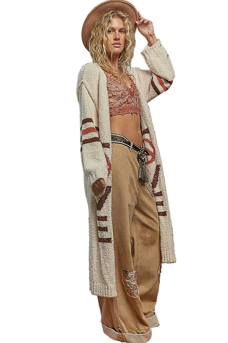 Woman wearing a long cardigan with patterned design, beige pants, and a hat on a white background