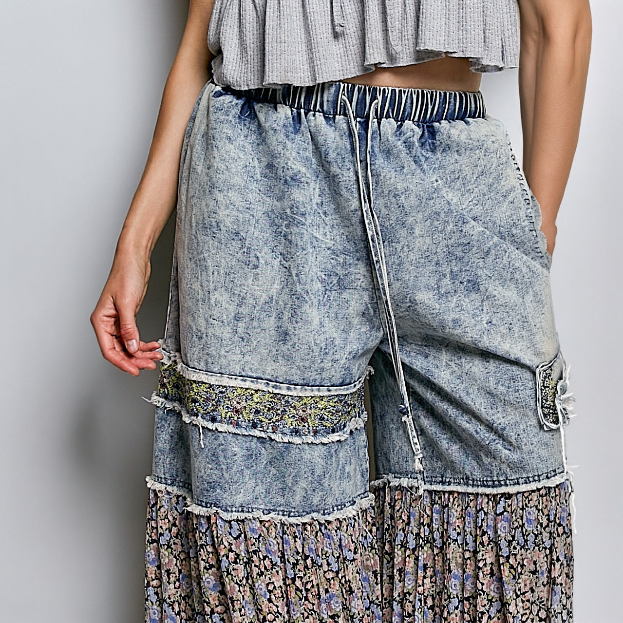 A close-up of a model wearing wide-leg, high-waisted denim shorts with a blue acid-wash finish, a striped elastic waistband, and a tiered floral skirted hem.