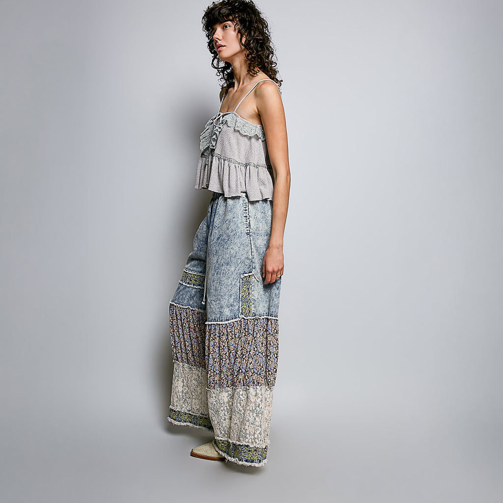 A woman models a two-piece bohemian-style outfit consisting of a light gray ruffled camisole top and wide-leg denim-wrapped pants with intricate patterned borders, set against a plain light gray studio background.