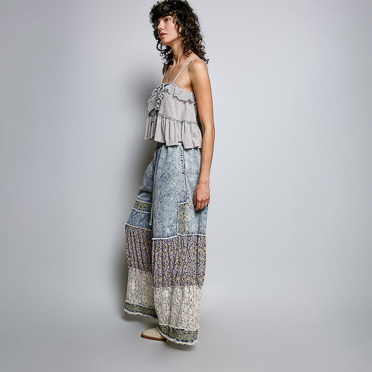 A woman models a two-piece bohemian-style outfit consisting of a light gray ruffled camisole top and wide-leg denim-wrapped pants with intricate patterned borders, set against a plain light gray studio background.