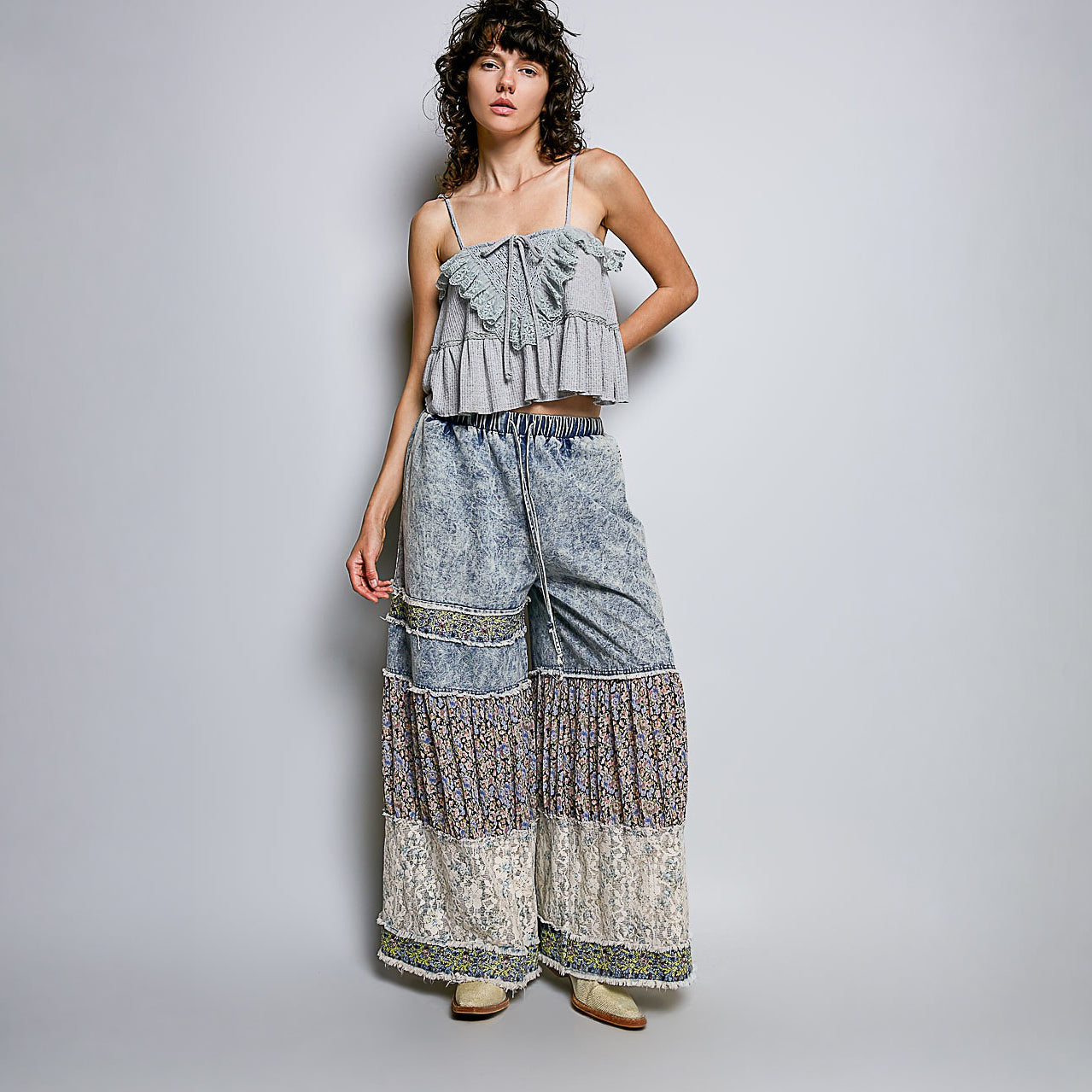 A model wearing a light gray ruffled camisole top and matching wide-leg patchwork pants with blue, beige, and brown patterns, standing against a light gray studio background.