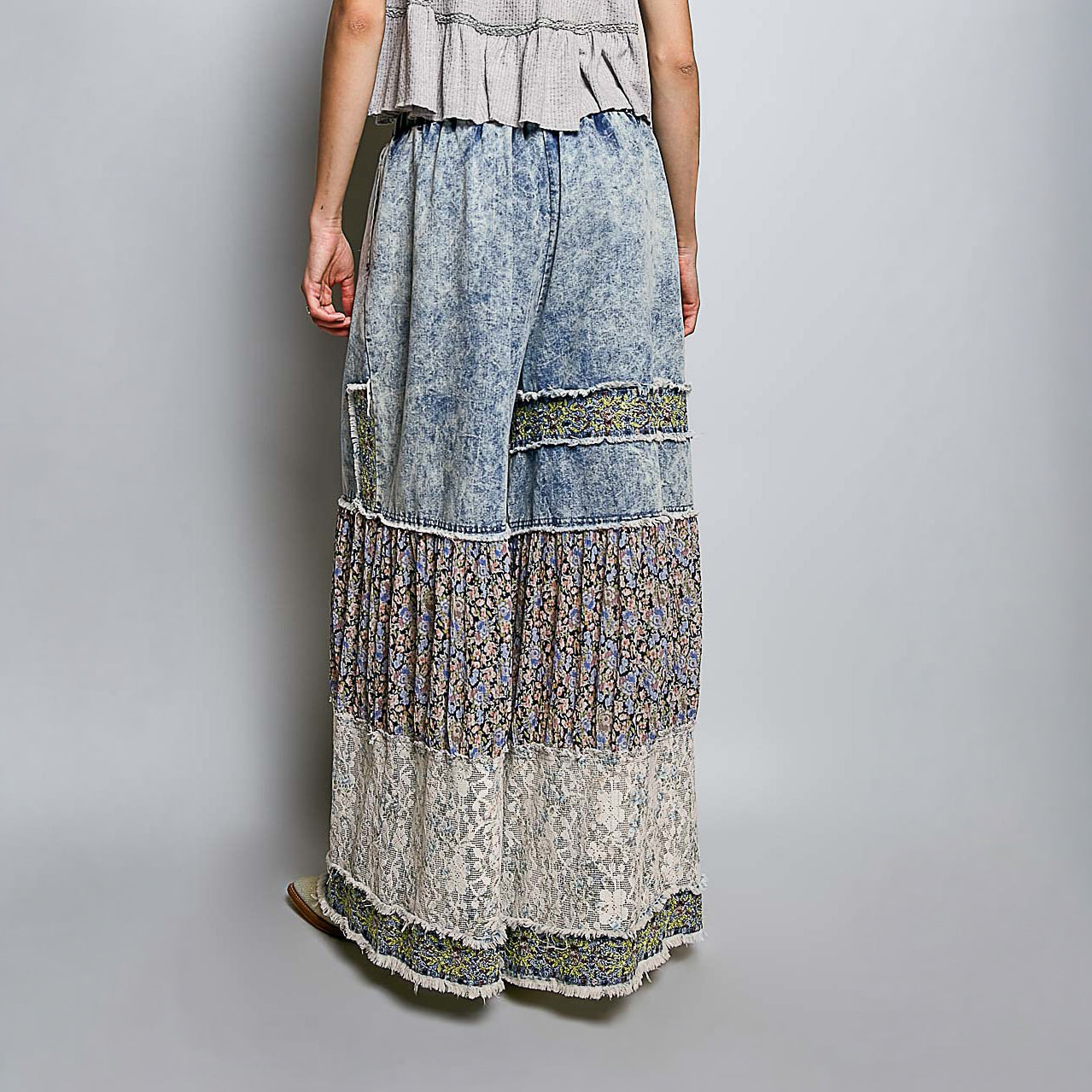 A model from the back wearing a long, tiered bohemian maxi skirt with a blue acid-wash denim top half and layered floral lace and patterned fabric panels below, set against a plain gray studio background.