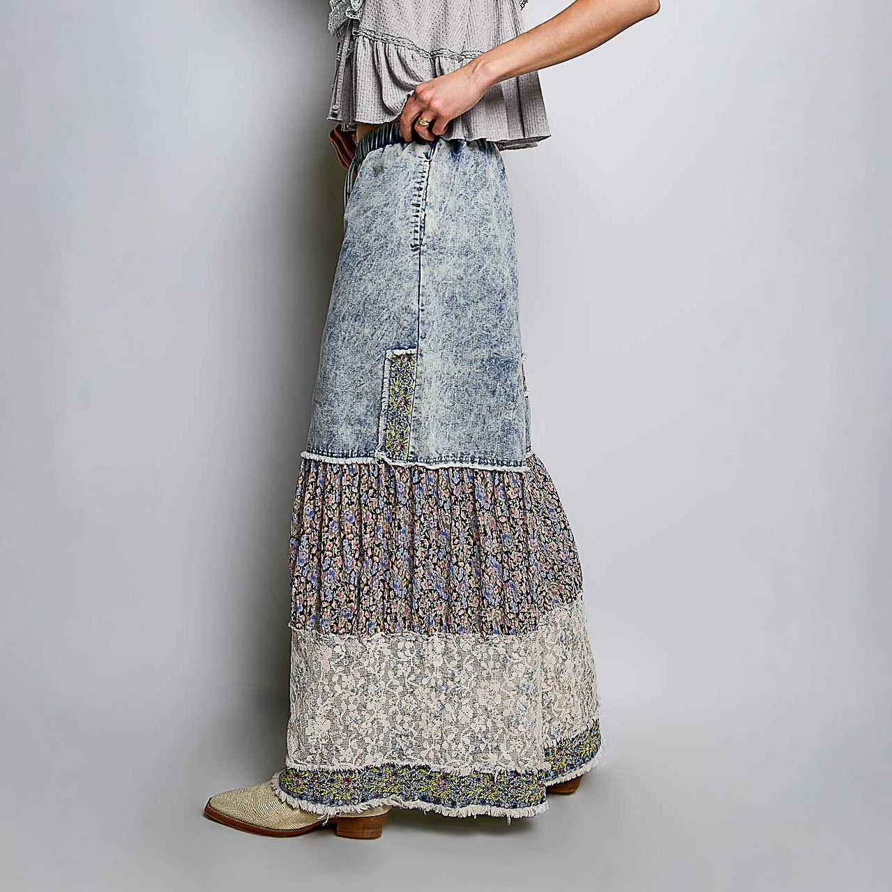 A model is shown from the waist down wearing a long, tiered maxi skirt with a light blue washed denim upper section and three layered ruffle sections in floral and lace patterns below, paired with beige boots against a neutral gray background.