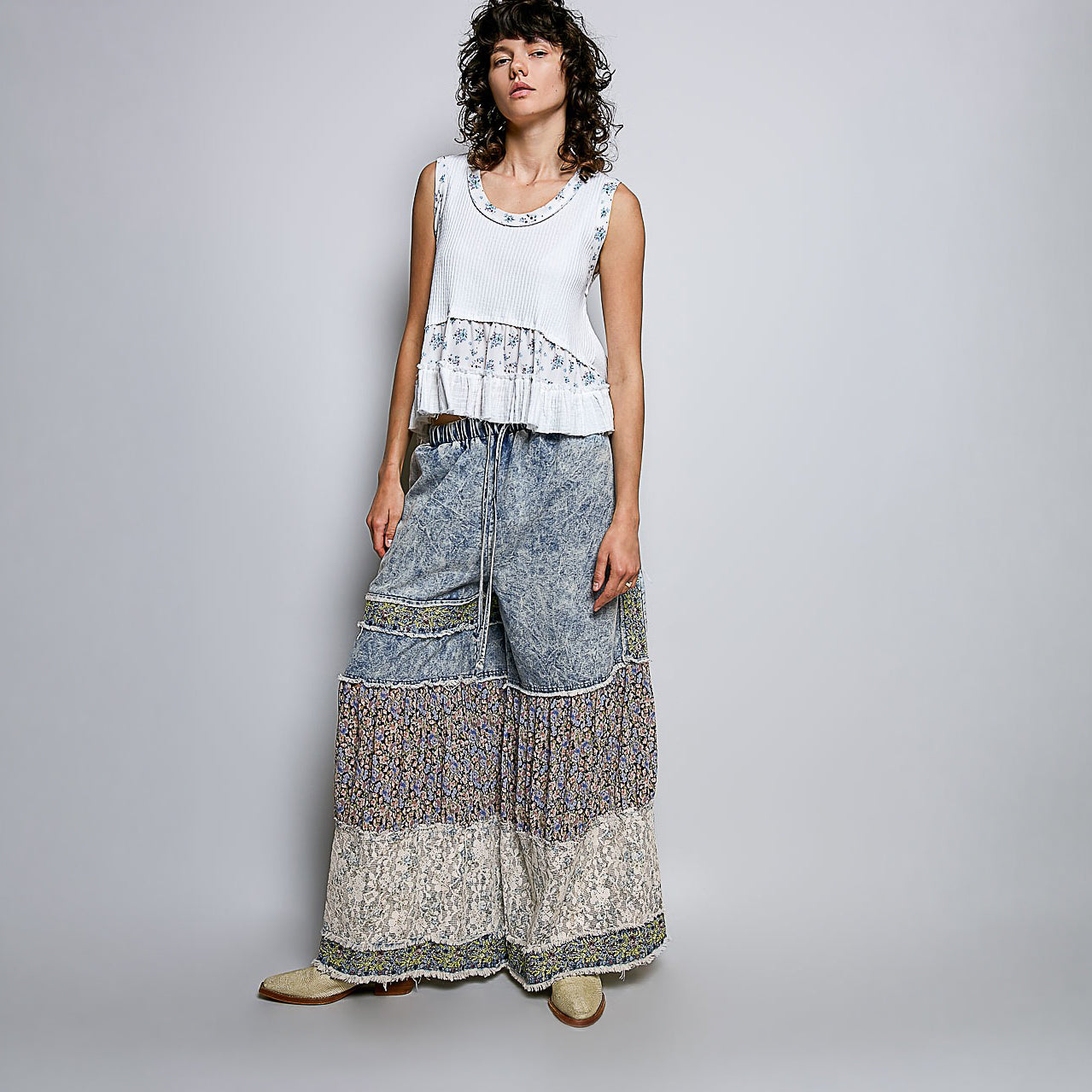 A woman modeling a sleeveless white top with a ruffled, patterned hem and matching wide-leg, patchwork-print pants against a light gray studio background.