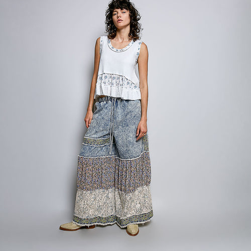 A woman modeling a sleeveless white top with a ruffled, patterned hem and matching wide-leg, patchwork-print pants against a light gray studio background.