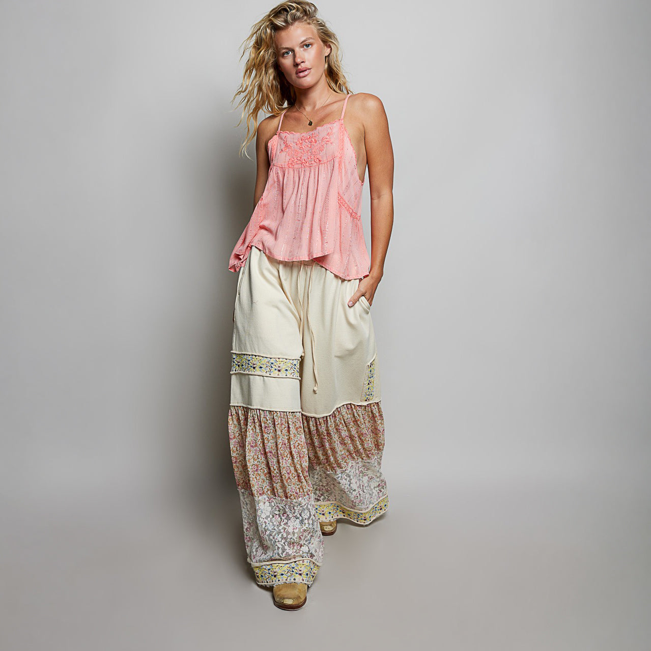 A woman modeling a bohemian-style two-piece outfit consisting of a light pink sleeveless top and cream wide-leg pants with tiered floral ruffle hems, set against a plain gray studio background.