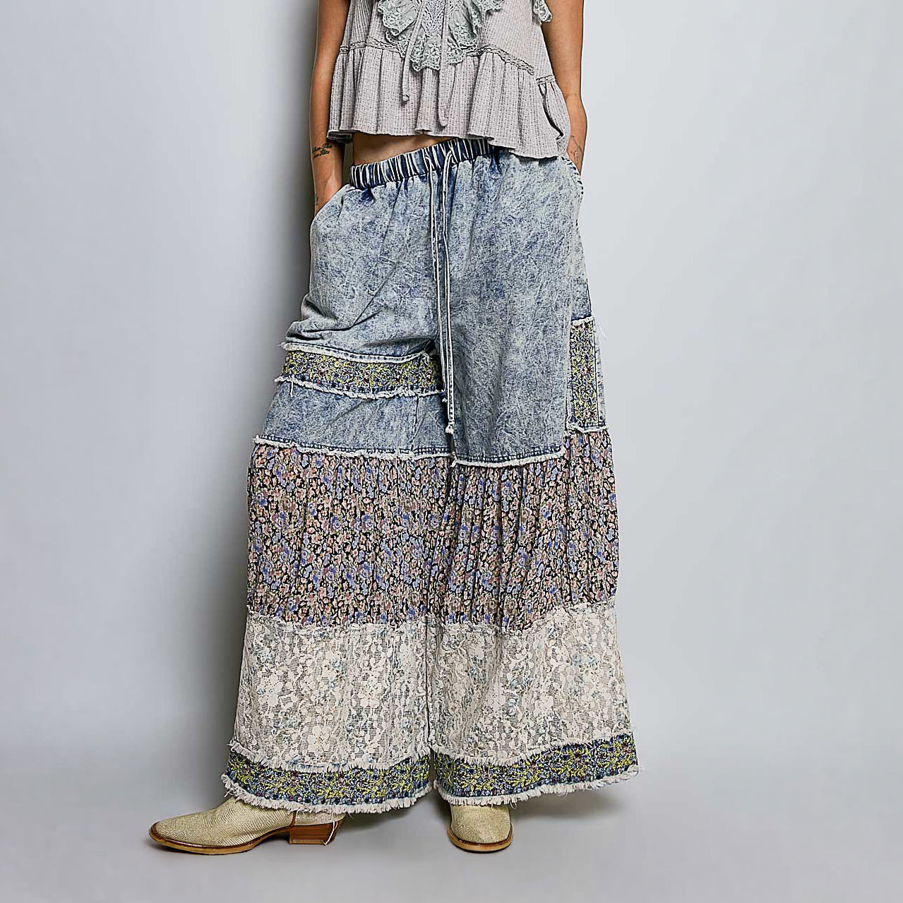 A women wearing wide-leg patchwork boho pants with layered floral and textured fabric panels in muted blue, beige, and gray tones, paired with a ruffled top and beige boots against a light gray studio background.