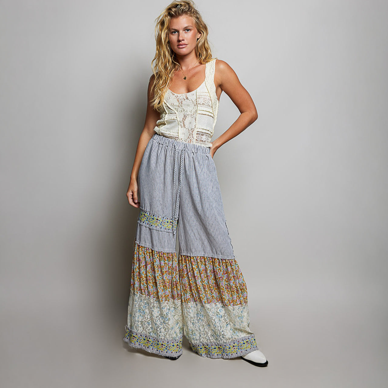 A woman modeling a sleeveless cream top and wide-leg striped maxi pants with multicolor floral and lace panels, photographed against a plain light gray studio background.