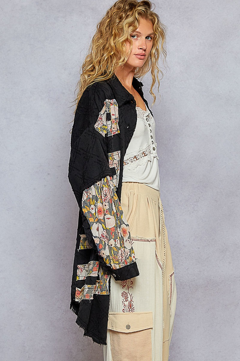 Woman wearing a black floral jacket over a white shirt with beige pants against a gray background