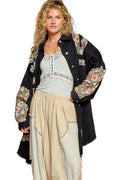 Woman wearing a black floral embroidered jacket over a light gray top and beige skirt on a white background