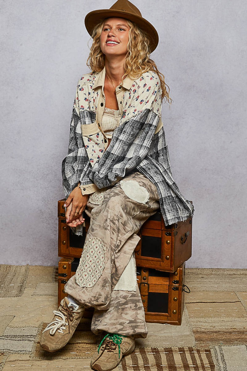 Woman sitting on a wooden suitcase wearing a plaid shirt, patterned pants, and a hat against a plain background.
