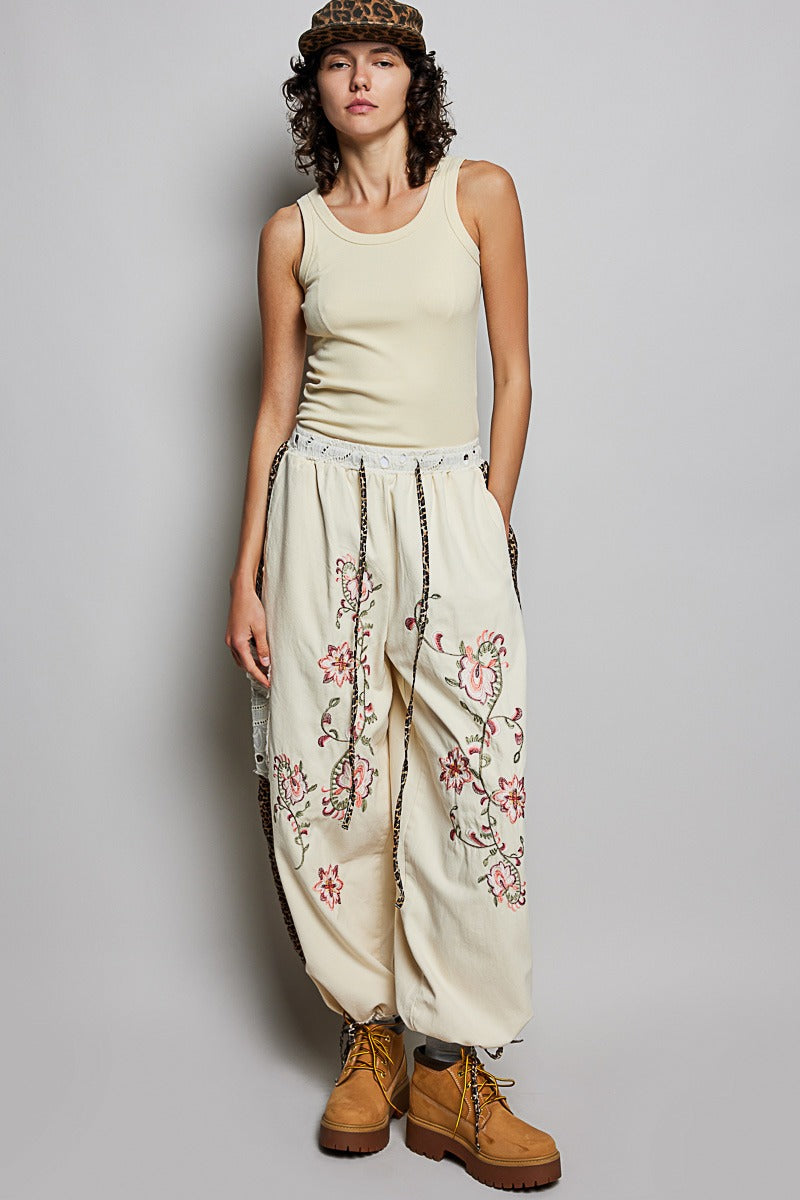 Woman wearing a beige tank top and floral embroidered pants against a plain background