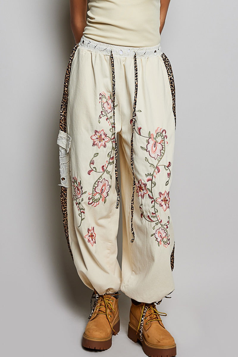 Beige pants with floral embroidery and leopard print trim worn by a person on a plain background