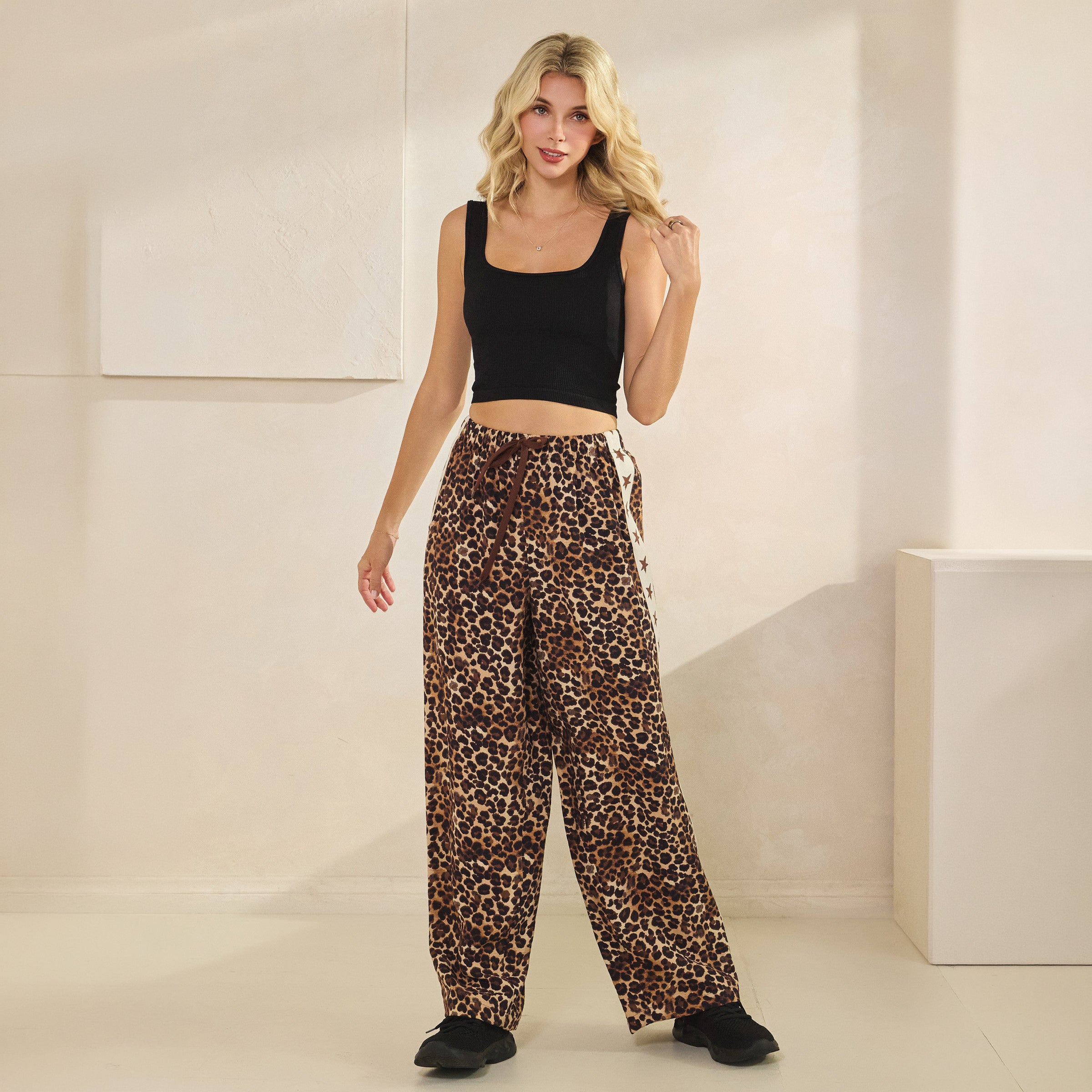 A woman modeling a black crop tank top and high-waisted leopard-print wide-leg pants with a side tie, standing in a minimalist beige room.