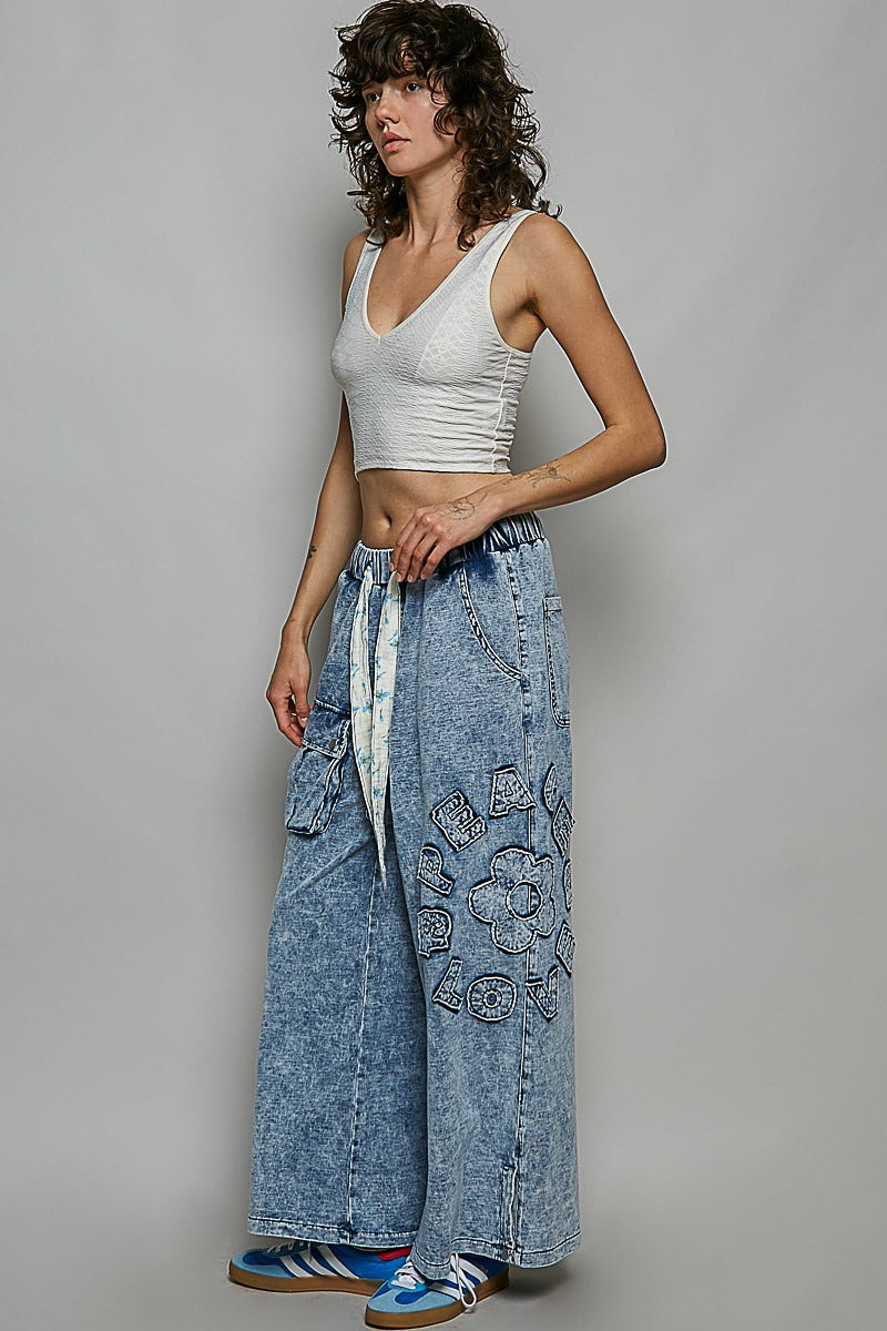 Woman wearing a white tank top and blue denim pants with floral patterns on a gray background