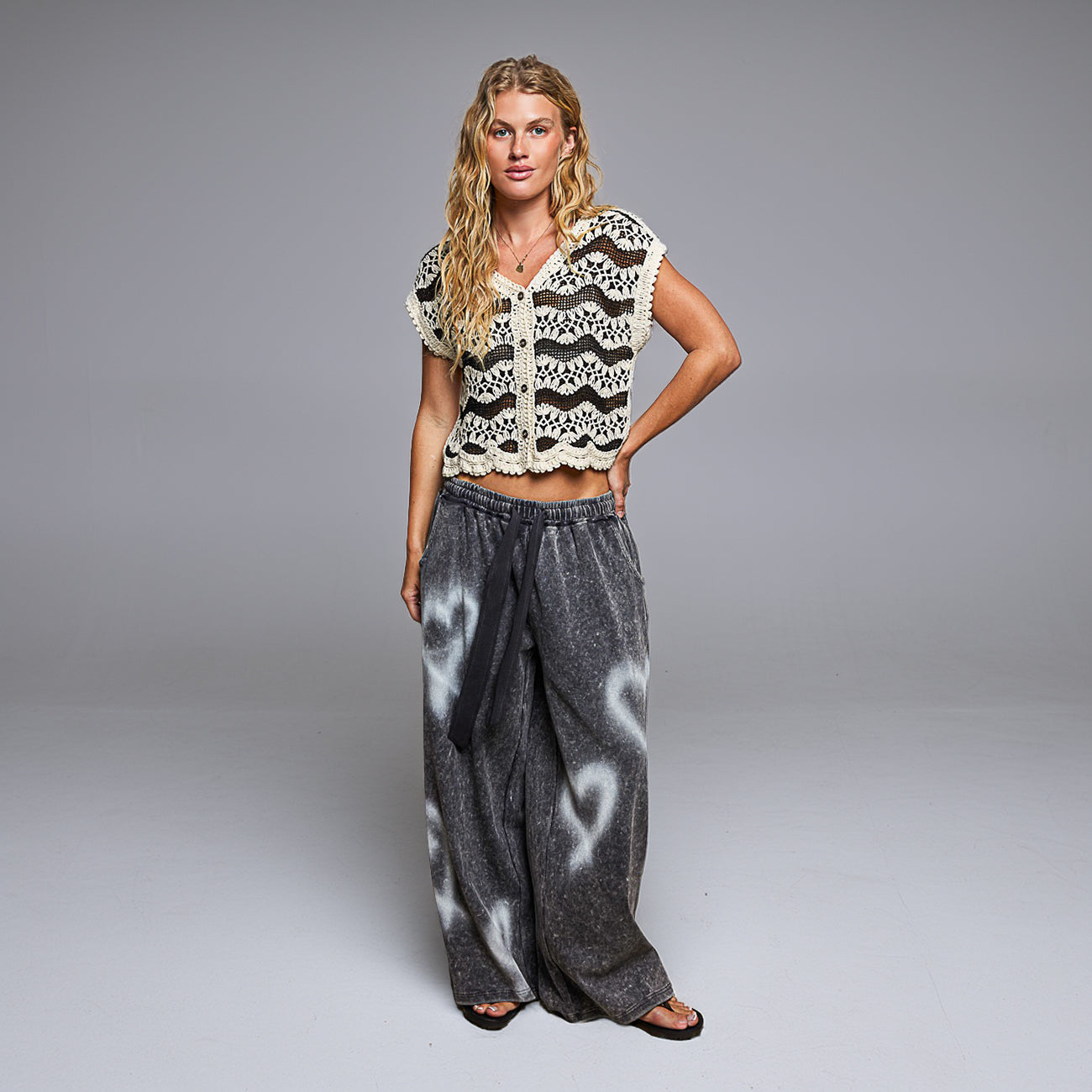 A woman modeling a short-sleeve crochet knit top with a black and cream chevron pattern and a matching cream lace trim, paired with wide-leg gray pants featuring white abstract patterns, against a solid gray studio background.