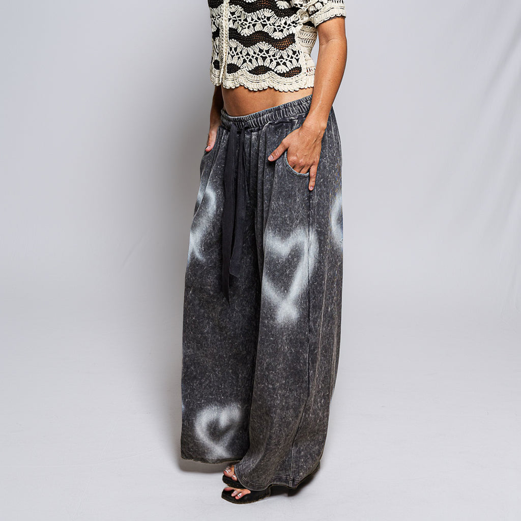 A person modeling wide-leg charcoal gray pants with a white heart spray-paint style pattern, paired with a black drawstring and black sandals, against a light gray studio background.