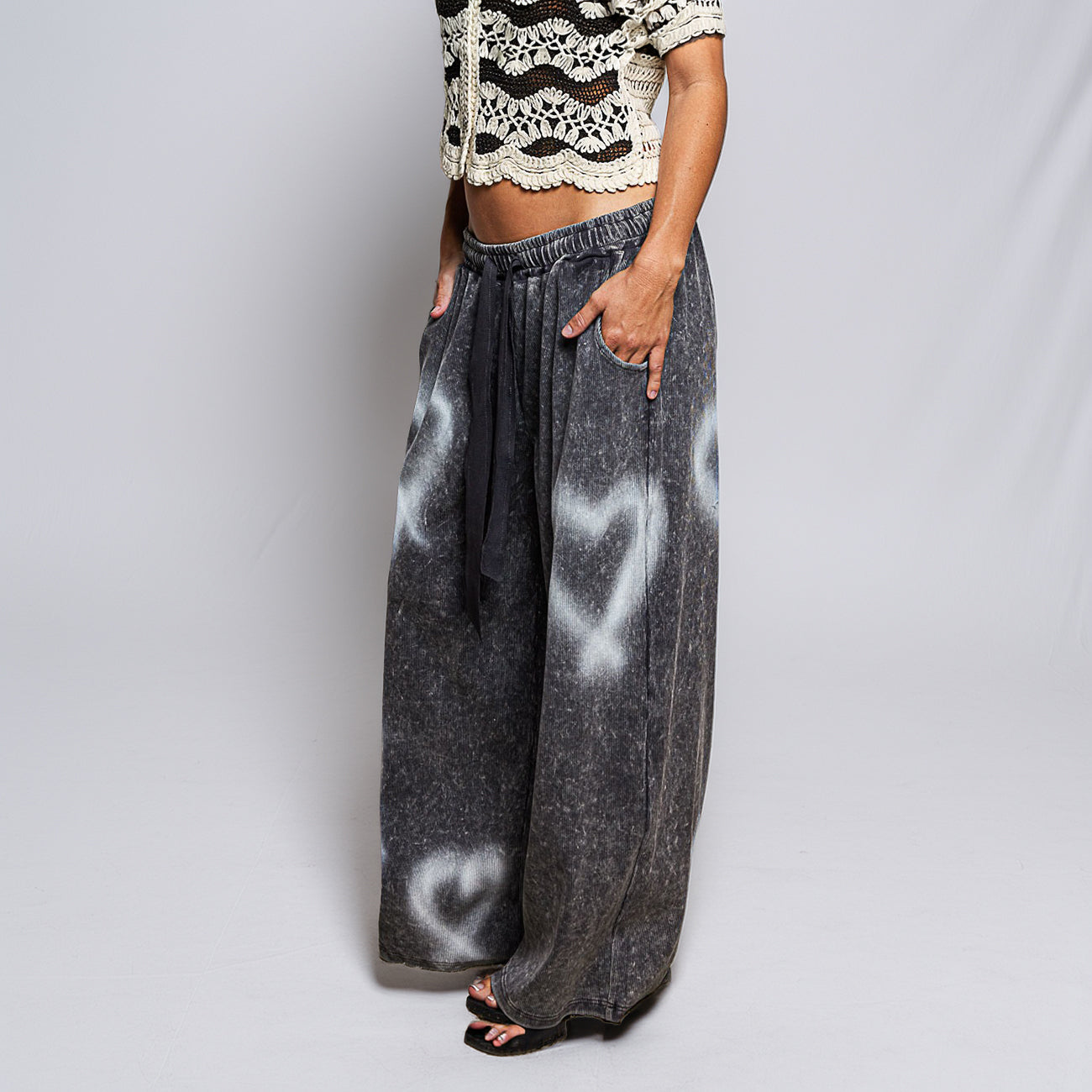 A person modeling wide-leg charcoal gray pants with a white heart spray-paint style pattern, paired with a black drawstring and black sandals, against a light gray studio background.