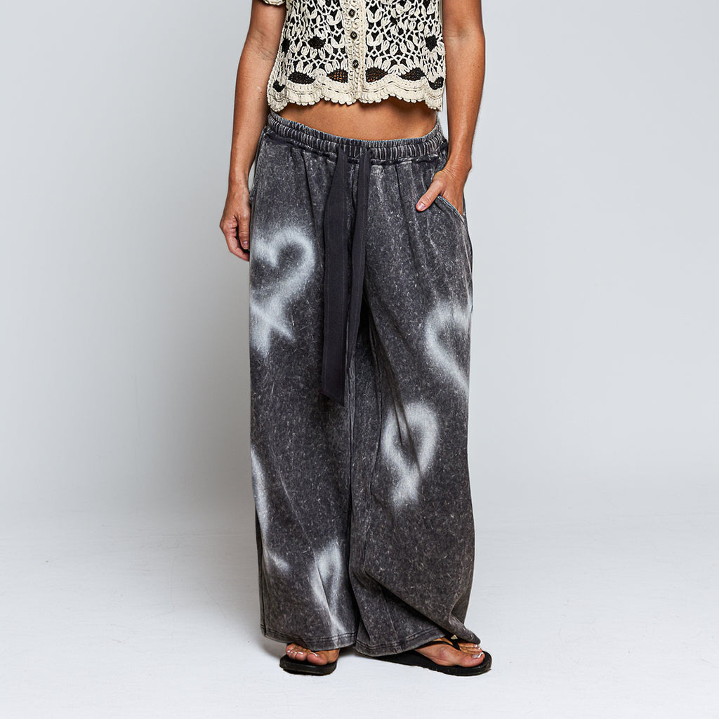 A person wearing wide-leg charcoal gray pants with white ribbon-like graphic prints and a black drawstring waist, paired with a cream crochet crop top and black sandals, against a light gray studio background.