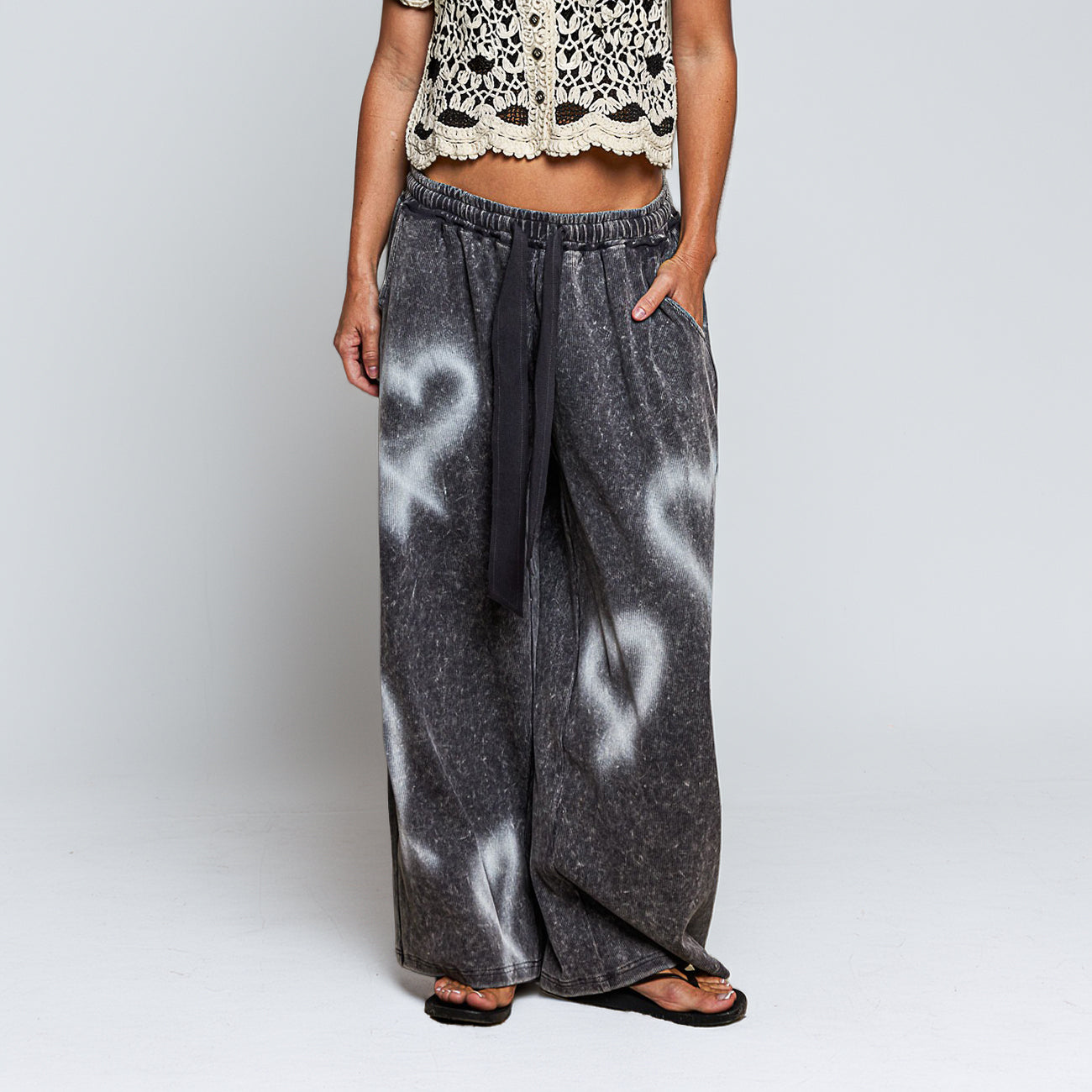 A person wearing wide-leg charcoal gray pants with white ribbon-like graphic prints and a black drawstring waist, paired with a cream crochet crop top and black sandals, against a light gray studio background.
