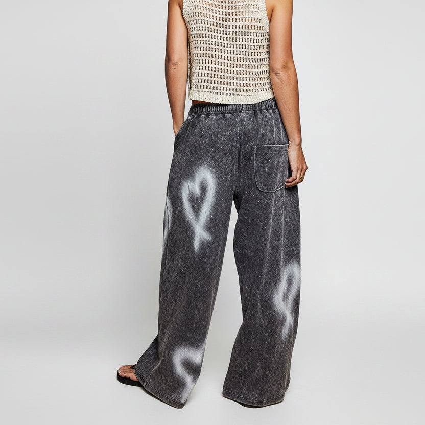 A person shown from the back wearing wide-leg charcoal gray acid-wash pants with white heart and ribbon motifs, paired with a cream crochet top against a plain white background.
