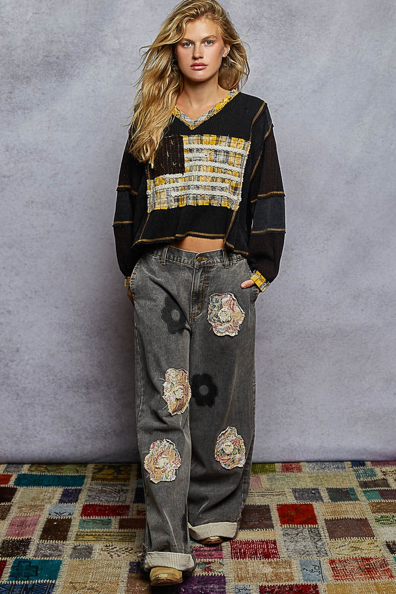 Woman wearing a black and gold top with floral patterns and gray pants with patches against a gray background.