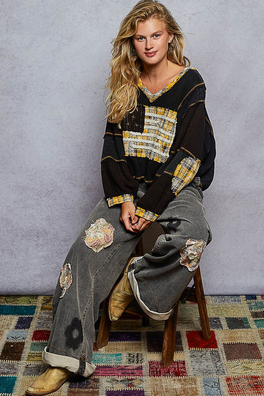 Woman wearing a black and yellow patterned top with gray pants sitting on a wooden stool.
