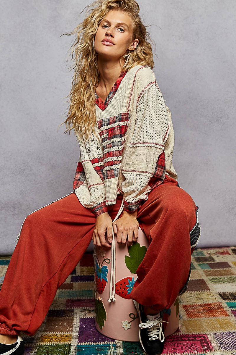Woman sitting on a colorful stool wearing a plaid shirt, beige cardigan, and red pants against a gray background.