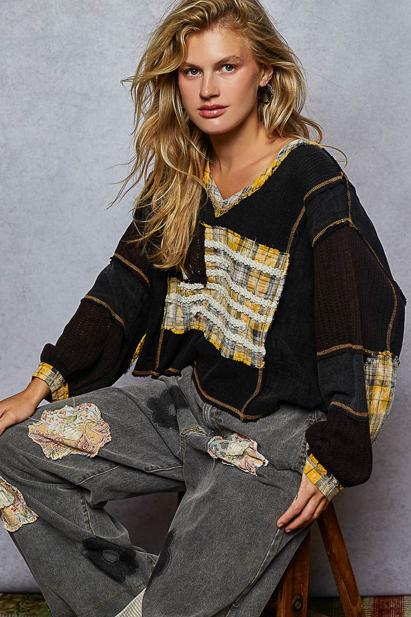 Woman wearing a black and yellow patterned top with gray pants, sitting on a stool against a gray background.