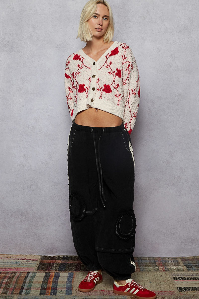 Person wearing a white cardigan with red floral patterns, black pants, and red sneakers against a gray wall.