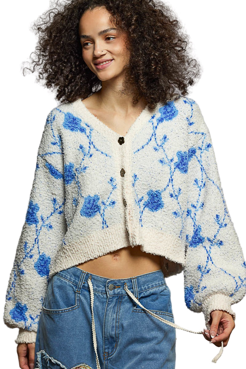 Woman wearing a white cardigan with blue floral patterns and blue jeans against a gray background