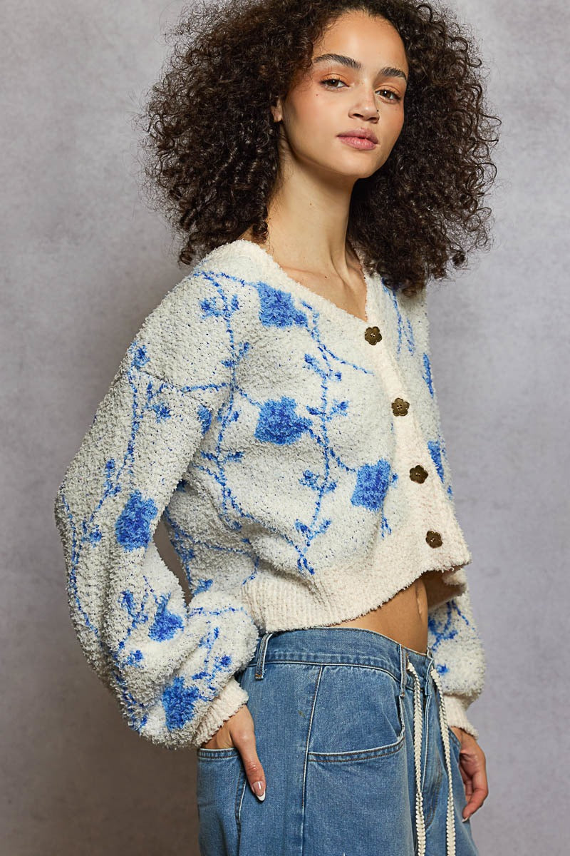 Woman wearing a white cardigan with blue floral patterns and blue jeans against a gray background