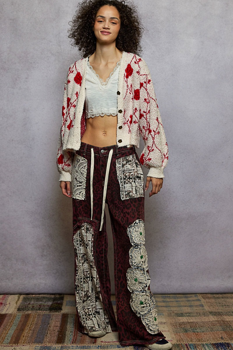 Woman wearing a patterned outfit with a white cardigan and brown pants against a gray wall.