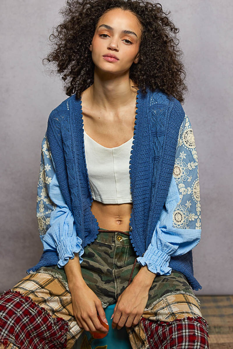 Woman wearing a blue cardigan over a white top with plaid pants against a gray background