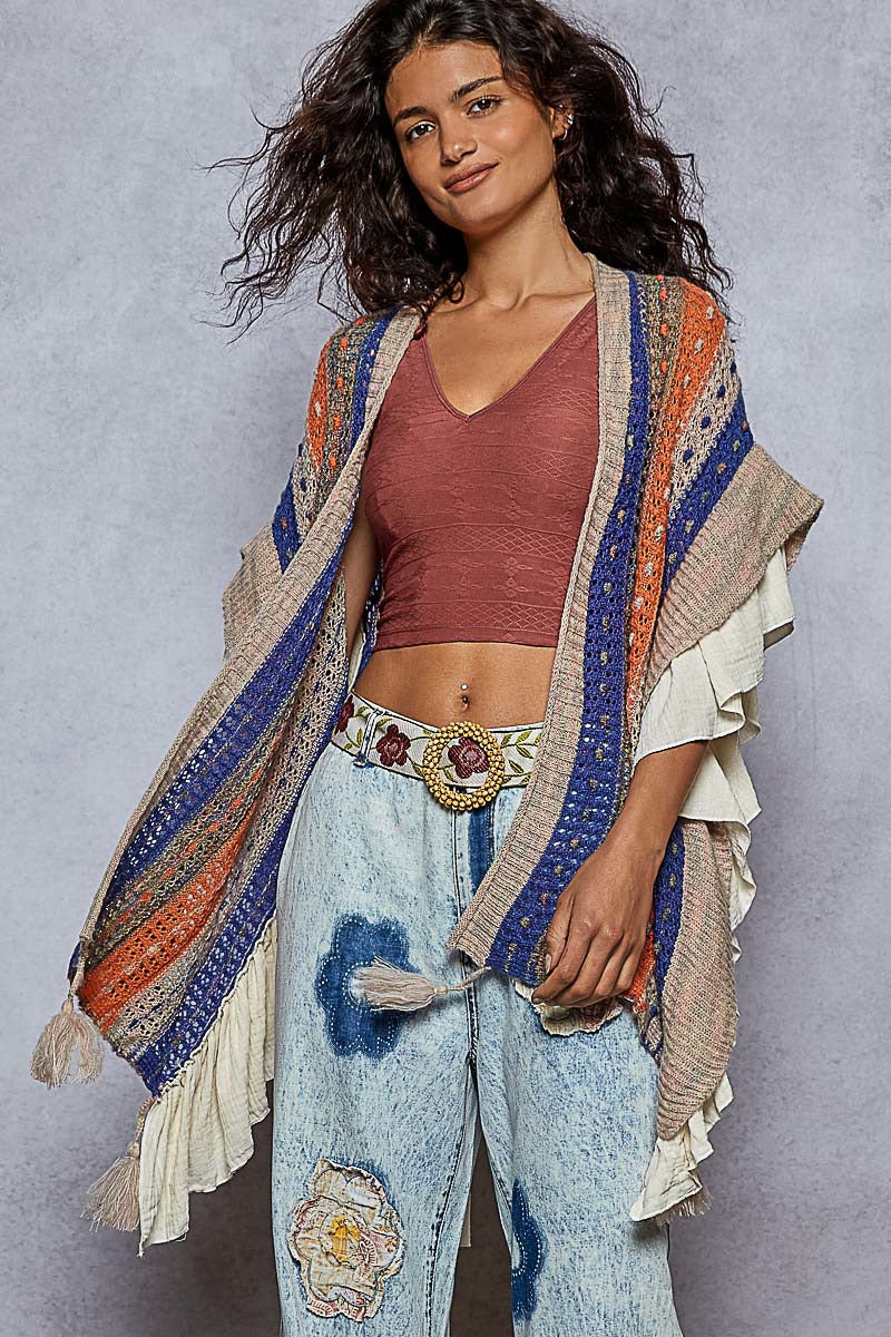 Woman wearing a colorful patterned shawl over a red top and light blue jeans with floral patches on a gray background