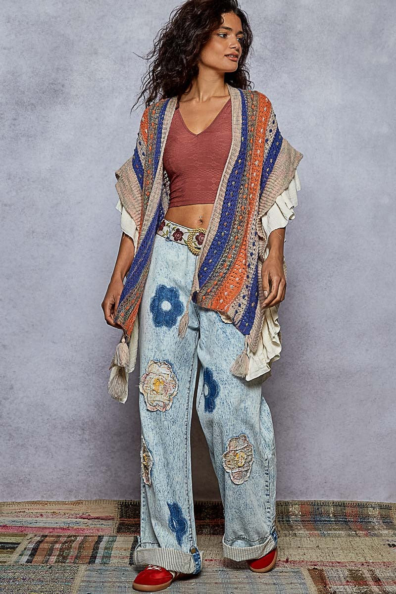 Woman wearing a colorful outfit with a patterned jacket and jeans against a gray wall.