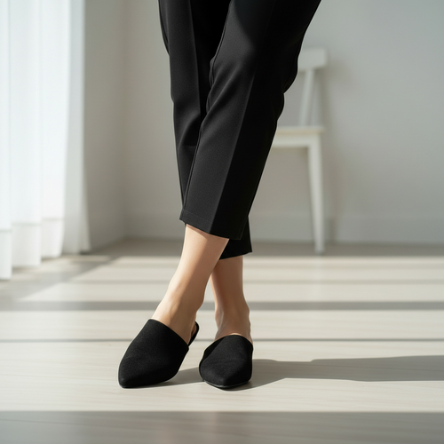 Person wearing black pants and black slippers standing on a light wooden floor.