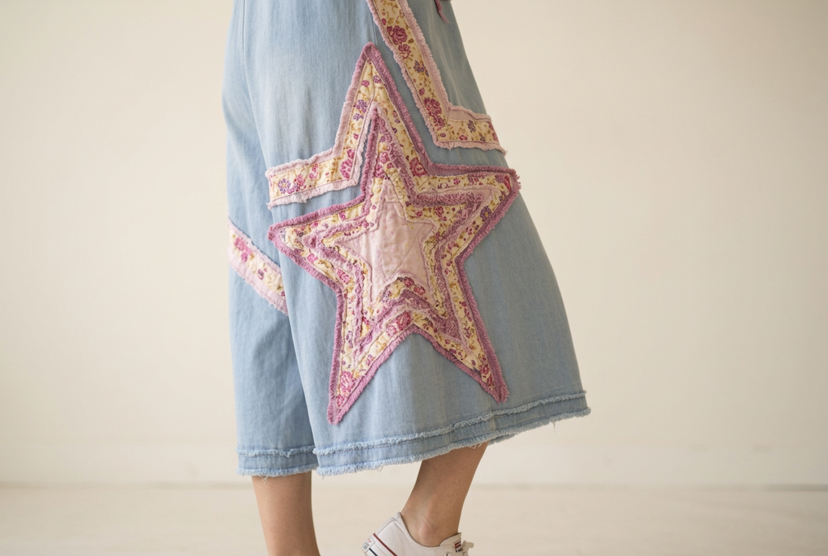 A close-up of a person wearing light blue denim shorts with a large embroidered star patch on the side in pink, yellow, and white, paired with white sneakers against a plain light beige background.