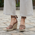Woman standing on stone pavement wearing taupe perforated suede espadrille wedge sandals with ankle-wrap ties, knees-down view