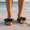 Close-up of feet wearing tropical print slide sandals on white sand beach with ocean waves