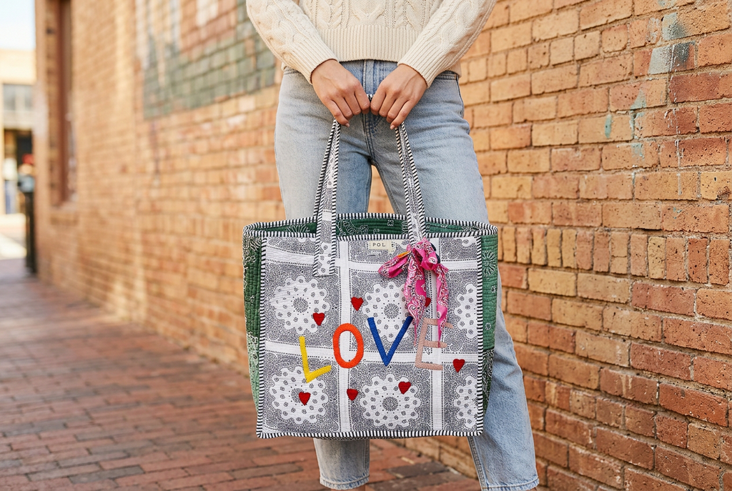Woman carrying a paisley LOVE tote with pink bandana beside a warm brick wall