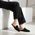 Woman seated on white bench wearing Bella Marie black pointed toe mule slides, outer side visible, ankles crossed, wide-leg trousers